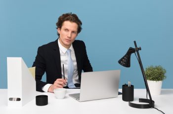 Homem vestido com esporte fino em frente a mesa com notebook pensando com uma caneta na mão