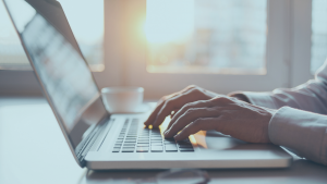 Mãos de um homem digitando em um notebook, com um pôr do sol ao fundo.