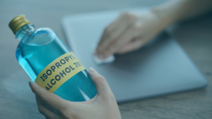 Pessoa segurando uma embalagem de álcool isopropílico, ao fundo usa um pano para limpar um notebook fechado.