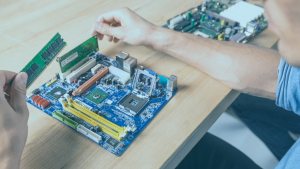 Placa eletrônica sobre uma mesa, nas mãos, uma pessoa segura a memória RAM