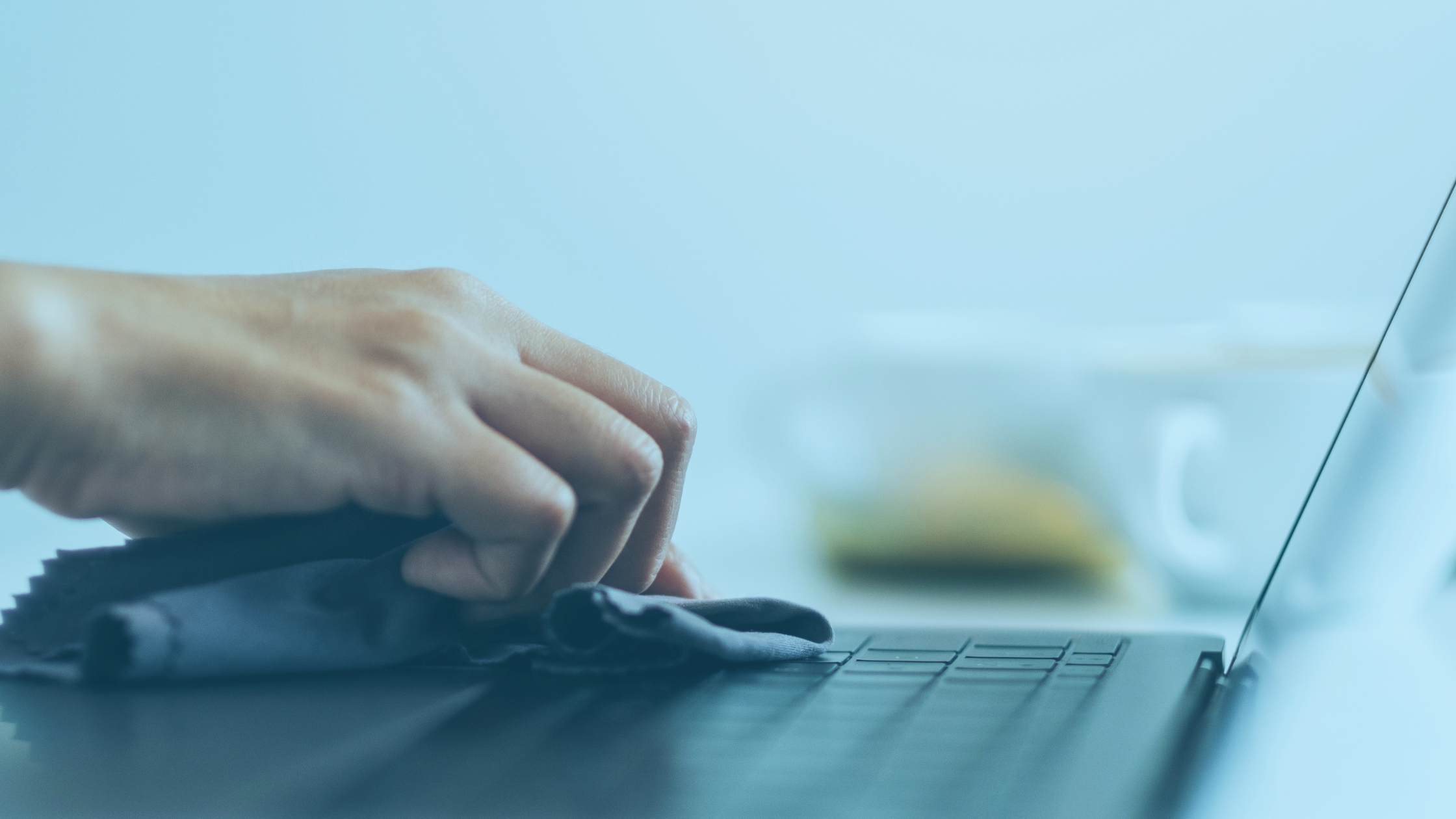 Visão lateral de um notebook, sobre o teclado há uma mão utilizando um pano e realizando a limpeza.