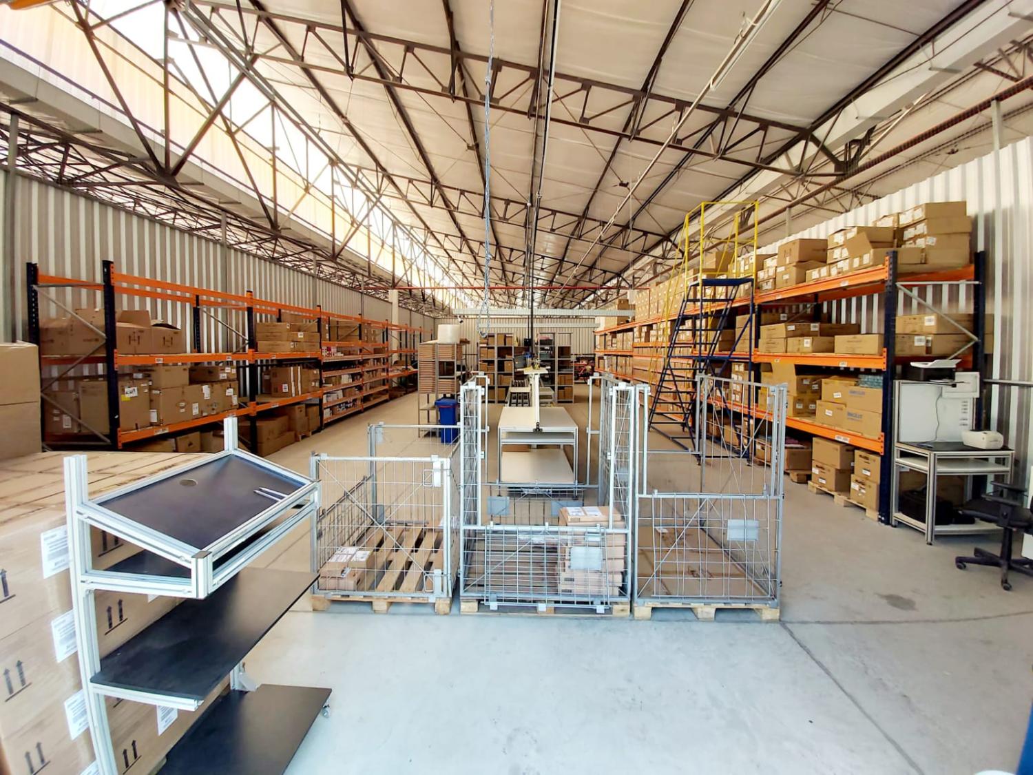 Centro de distribuição da bringIT em São Paulo, com prateleiras de metal laranja e azul organizadas com caixas e equipamentos em um grande galpão, mostrando a estrutura interna do armazém