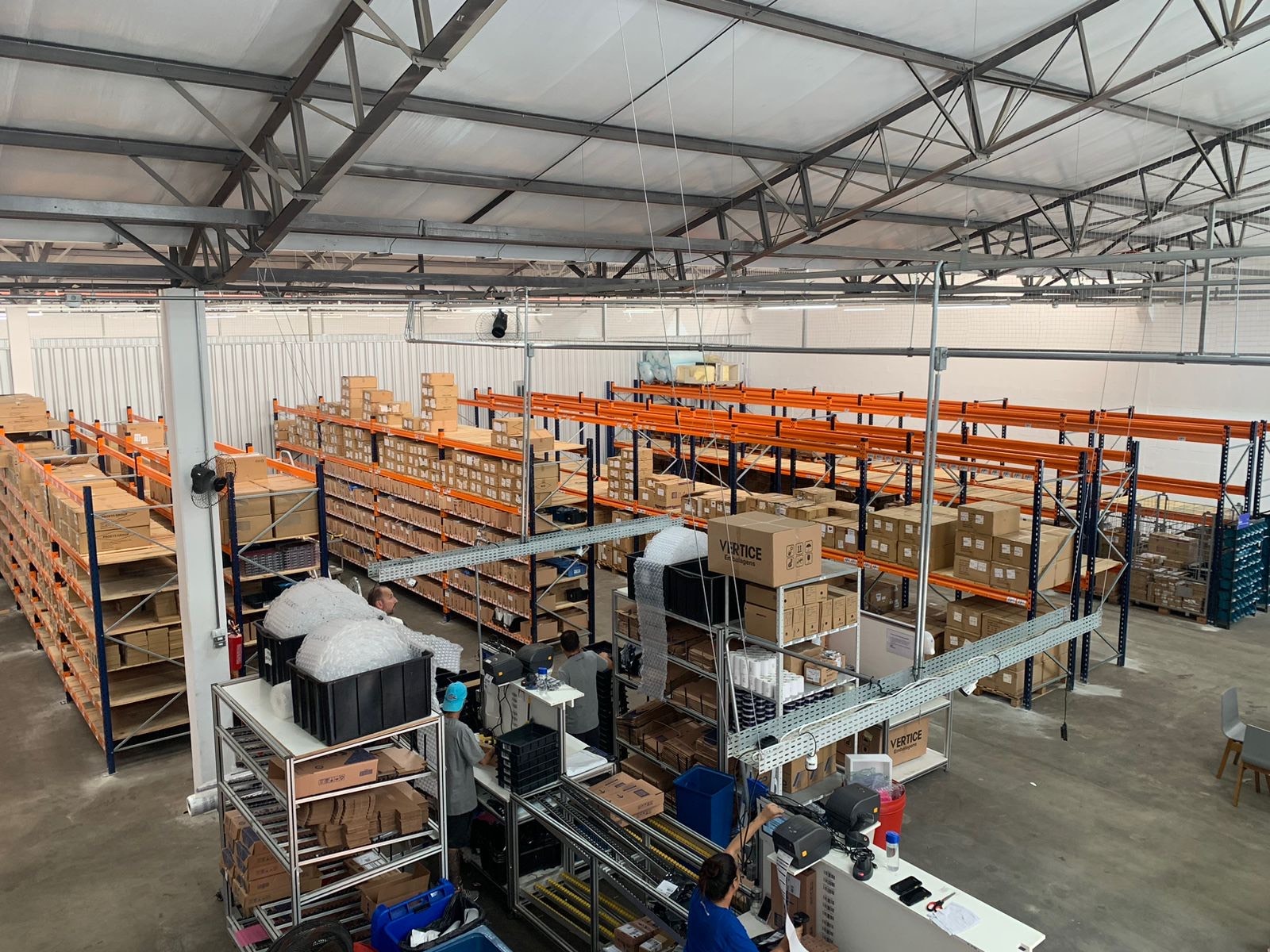 Centro de distribuição da bringIT em São Paulo, com prateleiras de metal laranja e azul organizadas com caixas e equipamentos em um grande galpão, mostrando a estrutura interna do armazém.