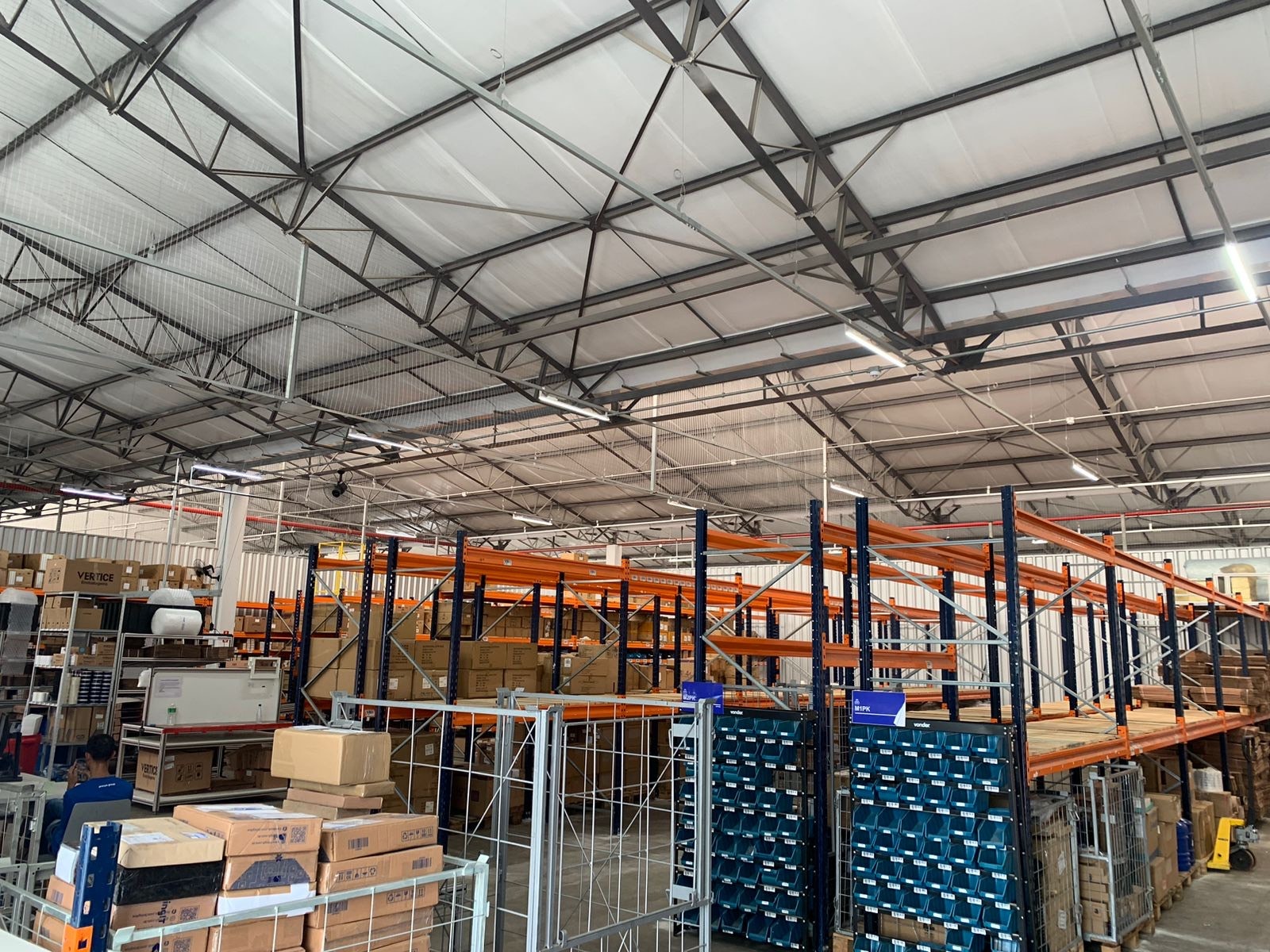 Centro de distribuição da bringIT em São Paulo, com prateleiras de metal laranja e azul organizadas com caixas e equipamentos em um grande galpão, mostrando a estrutura interna do armazém.